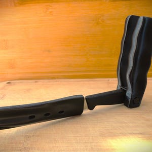 Può includere: Una maniglia nera stampata in 3D con un design curvo e un pezzo separato abbinato. La maniglia ha una superficie strutturata ed è progettata per essere fissata a un altro oggetto. Lo sfondo è una superficie di legno.