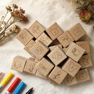 May include: Wooden alphabet blocks featuring engraved letters and illustrations, scattered on a cream-coloured surface. Markers are visible in the lower left corner, with dried flowers in the background. Each block is approximately 4 cm square.