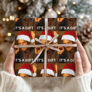 Peut inclure: Cadeau emballé avec un motif répété d'une personne portant un bonnet de Père Noël et le texte "IT'S A GIFT". Le cadeau est noué avec un ruban marron clair.
