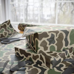 Op de afbeelding: Twee cadeau dozen verpakt in camouflage patroon papier, gestapeld op een tafel. Een rol bijpassend cadeaupapier en een schaar zijn zichtbaar op de achtergrond. Een grijs lint is op de tafel gekruld.
