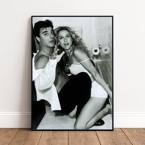 May include: Monochrome framed print showing a man and a woman in a bathroom. The man wears an open white shirt, the woman a white top and shorts. The image is a photograph, with a black frame.