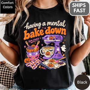 May include: A black t-shirt featuring a graphic with the text "having a mental bake down" and "from scratch". The design includes a purple stand mixer, mixing bowls, a baking sheet with cookies, ghosts, pumpkins, and bats in orange and purple.