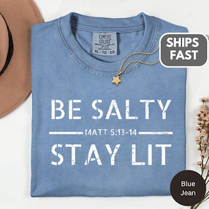 以下が含まれることがあります： 「BE SALTY」と「STAY LIT」の白い文字がプリントされた、ブルーデニムのComfort Colors Tシャツ。「MATT 5:13-14」の文字もプリントされています。金の星のペンダントネックレスがTシャツの上にあります。