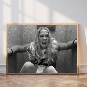 May include: Black and white framed print featuring a person in a distressed pose. The person is wearing a white t-shirt with a floral design and the word "Lou" printed on it, a red jacket, and white underwear. The background has a floral wallpaper design.