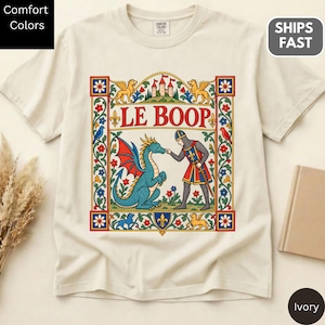 May include: An ivory short-sleeve t-shirt featuring a central graphic of a knight and a blue dragon with red wings. The text LE BOOP is printed above the figures, surrounded by a decorative border with floral motifs, lions, and a castle.