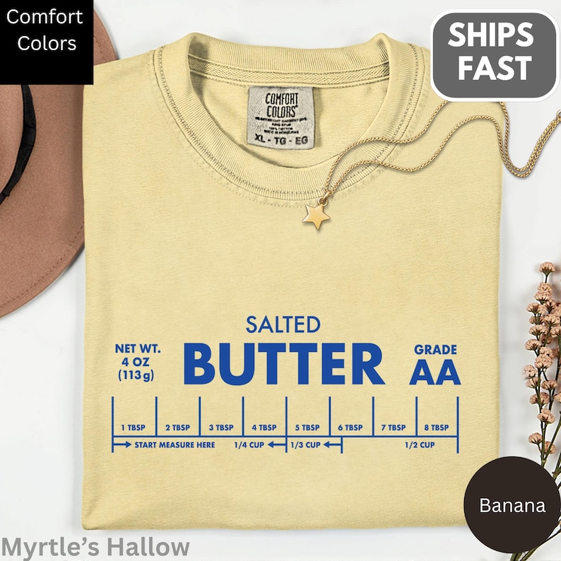 a yellow t-shirt with a blue text design that reads "SALTED BUTTER AA" and a chart showing the measurements of butter in cups and ounces. The t-shirt is displayed on a white background, and there is a small plant and a hat to the left of the t-shirt.