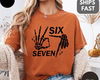 以下が含まれることがあります： 「SIX SEVEN」の文字と骸骨の手のグラフィックがプリントされた、オレンジ色のTシャツ。 Comfort Colorsブランドで、ゆったりとしたフィット感。 デザインは黒で、平和のサインをする骸骨の手が特徴です。