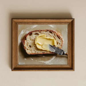 Op de afbeelding: Een sneetje brood met boter dat er met een mes op wordt gesmeerd. Het brood zit in een houten frame en de boter heeft een lichtgele kleur. Het mes heeft een zilveren lemmet en een houten handvat.