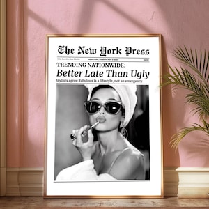 以下が含まれることがあります： 新聞の表紙を額装した白黒プリント。見出しは「Better Late Than Ugly」で、女性が口紅を塗っています。新聞のタイトルは「The New York Press」です。プリントは金のフレームに入っています。
