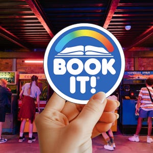 May include: A blue, white, and rainbow circular sticker with the text "BOOK IT!" is held in front of a retro arcade. The sticker features an open book design with a rainbow arch. The arcade has neon lights and people playing games.