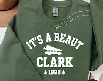 Może przedstawiać: Zielona wojskowa bluza z białym napisem "IT'S A BEAUT CLARK 1989". Projekt zawiera samochód z choinką. Bluza ma okrągły dekolt.