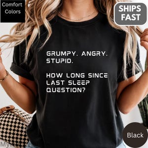 Op de afbeelding: Zwart T-shirt met witte tekst: "GRUMPY. ANGRY. STUPID. HOW LONG SINCE LAST SLEEP QUESTION?" Het shirt heeft een ronde hals en korte mouwen. De woorden "Comfort Colors" en "SHIPS FAST" zijn ook zichtbaar.