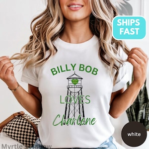 May include: White t-shirt with the text "BILLY BOB LOVES Charlene" in green, with a black and green water tower graphic. The shirt has short sleeves and a crew neck.