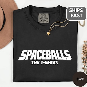 May include: Black t-shirt with the text "SPACEBALLS THE T-SHIRT" in white block letters. The shirt has a crew neck and short sleeves. A gold-colored star pendant hangs from a delicate chain. The shirt is labeled XL-TG-EG.
