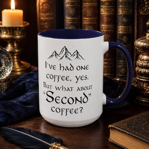 May include: A white ceramic mug with a dark blue interior and handle. The front features a black mountain graphic and the text: I've had one coffee, yes. But what about "Second" coffee? The mug has a cylindrical body and a curved handle.