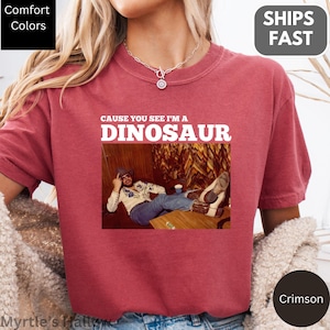 a person wearing a red t-shirt with a graphic design featuring a person lying down and the text "CAUSE YOU SEE I'M A DINOSAUR" printed on it.