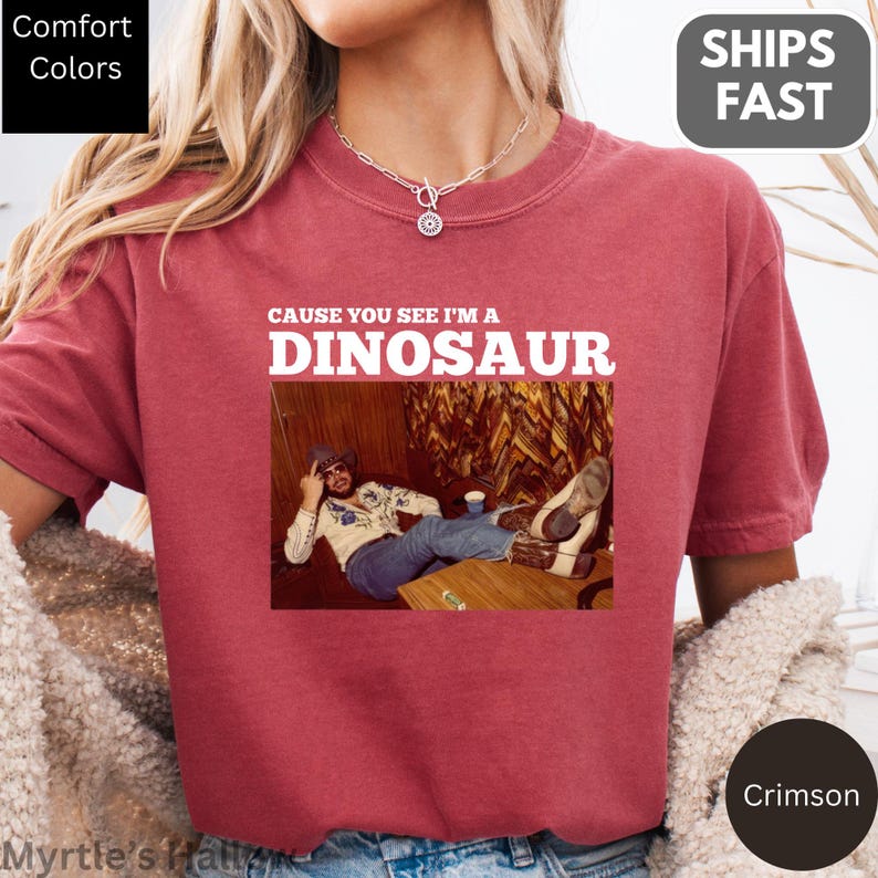 a person wearing a red t-shirt with a graphic design featuring a person lying down and the text "CAUSE YOU SEE I'M A DINOSAUR" printed on it.