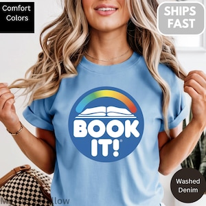 Könnte beinhalten: Hellblaues T-Shirt mit einem kreisförmigen Aufdruck, der ein offenes Buch unter einem Regenbogenbogen und die Worte "BOOK IT!" zeigt. Das T-Shirt hat kurze Ärmel. Die Texte "Comfort Colors" und "Washed Denim" sind ebenfalls sichtbar.