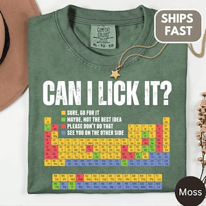 Peut inclure: Un t-shirt vert mousse avec le texte blanc CAN I LICK IT? au-dessus d'un tableau périodique coloré. Le graphique comprend une légende avec des carrés jaunes, verts, rouges et bleus indiquant les niveaux de sécurité. Un collier étoile doré est posé sur le col.