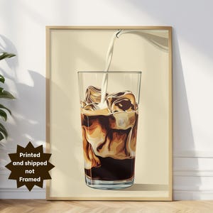 May include: A framed print of a glass of iced coffee with milk being poured in. The coffee is dark brown, and the milk is white, creating a swirl effect. The glass is filled with ice cubes. The background is a light beige color.