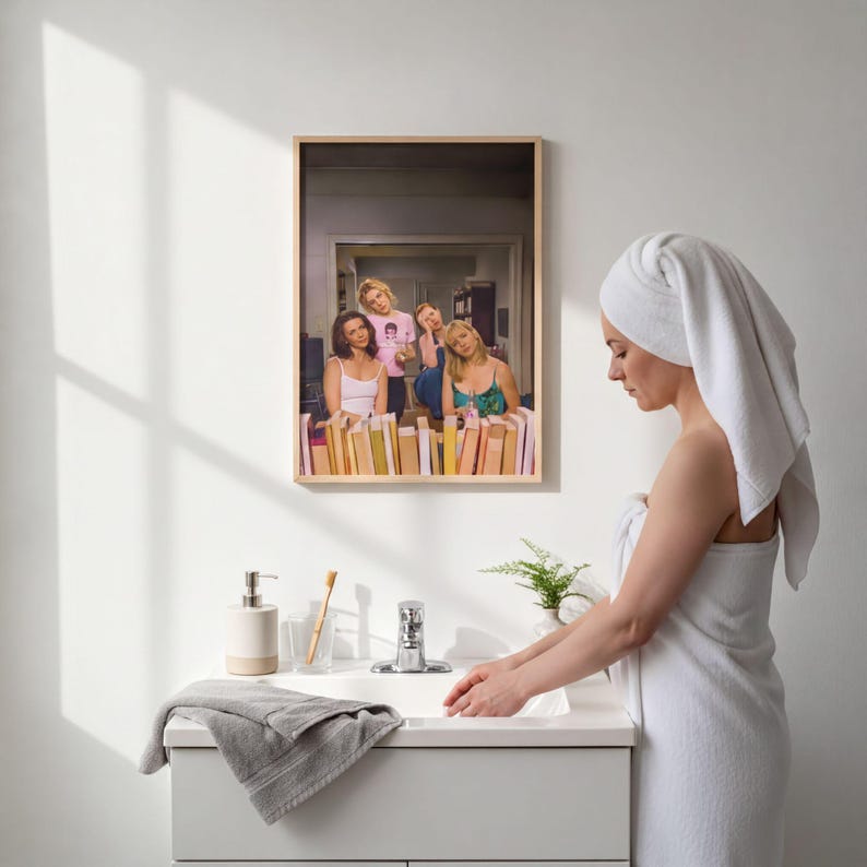 A woman in a white towel stands at a sink in a bathroom, with a framed photo of three people on the wall behind her.