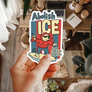 以下が含まれることがあります： 太字で「Abolish ICE」という言葉が入ったステッカー。赤いパーカーとサングラスをかけた漫画のキャラクターがスプレー缶を持っています。「keep families together」というテキストも含まれています。ステッカーは手で持たれています。