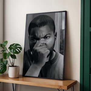 A framed black and white photograph of a man with a serious expression is displayed on a wooden shelf next to a potted plant.