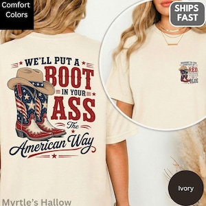 May include: Ivory-colored t-shirt with a graphic on the back featuring a cowboy hat and boots in red, white, and blue, with the text "WE'LL PUT A BOOT IN YOUR ASS The American Way." A smaller graphic is on the front.