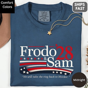 Op de afbeelding: Een marineblauw t-shirt met korte mouwen voorzien van de wit-rode tekst Frodo & Sam '28, een afbeelding van een wapperende Amerikaanse vlag onder de tekst en de zin We will take the ring back to Mordor in het wit onderaan gedrukt.