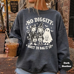 Puede incluir: Una sudadera de cuello redondo en gris jaspeado oscuro con un estampado gráfico blanco de tres niños con disfraces de Halloween, incluyendo un diablo, una bruja y un fantasma, cada uno con un cubo de calabaza, con el texto NO DIGGITY BOUT TO BAG IT UP.