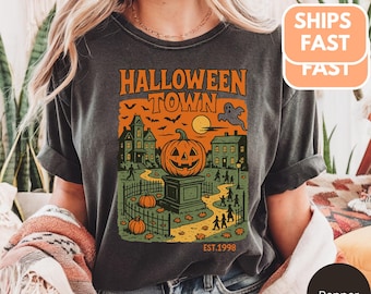 May include: Dark gray t-shirt with a Halloween-themed graphic. The design features a jack-o'-lantern, haunted houses, and the text "HALLOWEEN TOWN EST.1998" in orange and green. The shirt is a relaxed fit.
