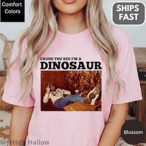 A woman wearing a pink t-shirt with a graphic design featuring a man lying down and the text "CAUSE YOU SEE I'M A DINOSAUR".