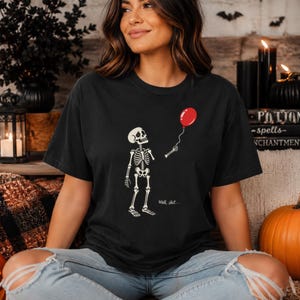 Può includere: Una t-shirt nera con una grafica bianca di uno scheletro che tiene in mano un palloncino rosso. Lo scheletro guarda verso il palloncino, che è staccato dalla sua mano. Il testo "Well, shit..." è stampato in piccole lettere bianche sotto.