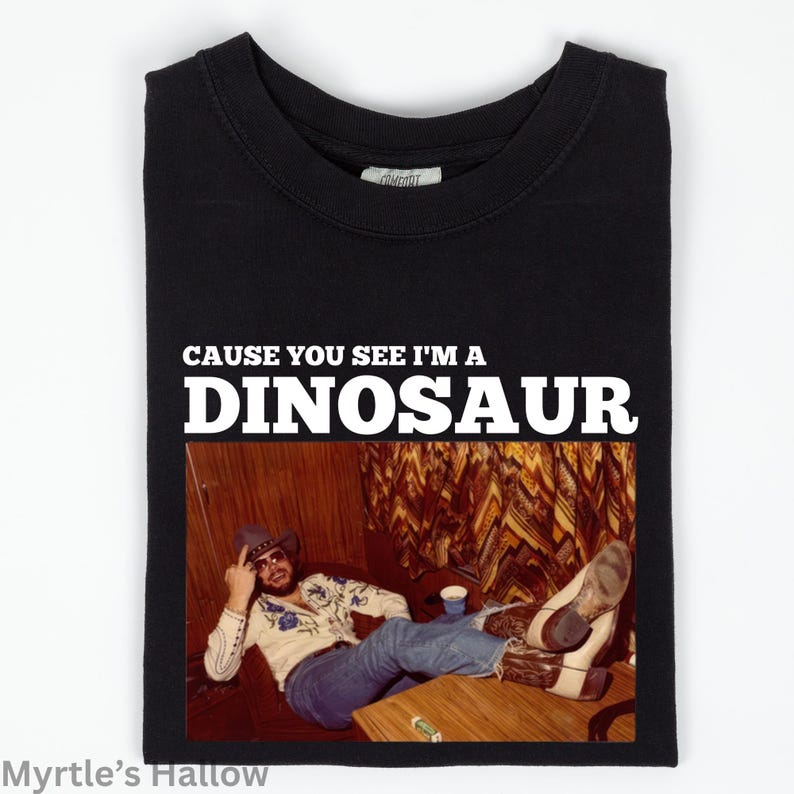 The image is a black t-shirt with a printed design that includes a man sitting on a couch and the text "CAUSE YOU SEE I'M A DINOSAUR" above the image.