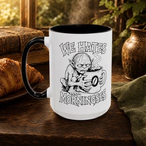 May include: A white ceramic mug with a black interior and handle, featuring a black line art illustration of a creature holding a coffee cup. The text "WE HATES MORNINGSES" is printed in black above and below the creature, with a coffee bean icon.