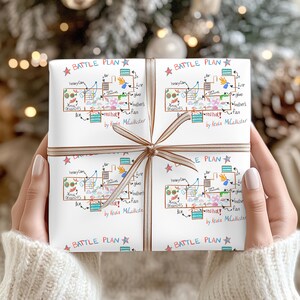 May include: White gift wrap with a colourful "Battle Plan" design, featuring hand-drawn illustrations and text. The wrap is tied with a brown ribbon. Text includes "heavy Cam", "Xmas ornaments", "ice", "fire", "glue", and "red hot!"