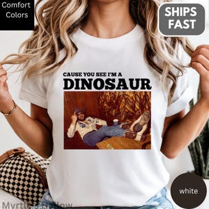 A person wearing a white t-shirt with a graphic design featuring a man sitting on a wooden bench with a dinosaur-like figure in the background.