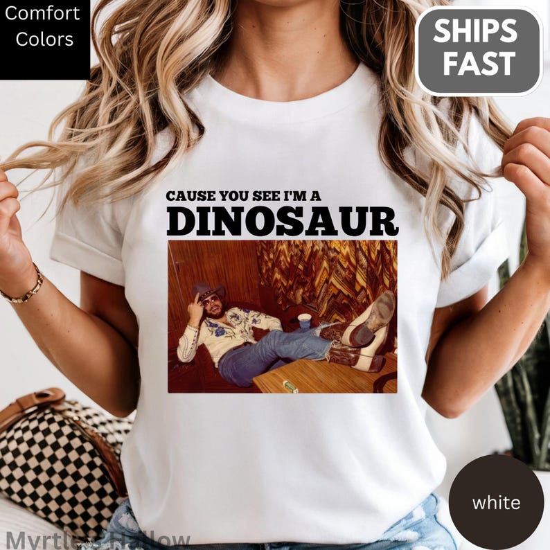 A person wearing a white t-shirt with a graphic design featuring a man sitting on a wooden bench with a dinosaur-like figure in the background.