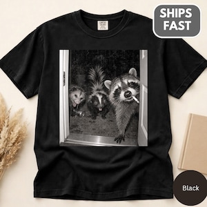 Puede incluir: Una camiseta negra de manga corta con un estampado gráfico de un mapache, una mofeta y una zarigüeya en una puerta. El mapache está en primer plano con un cigarrillo en la boca. La camiseta tiene una etiqueta de cuello Comfort Colors.