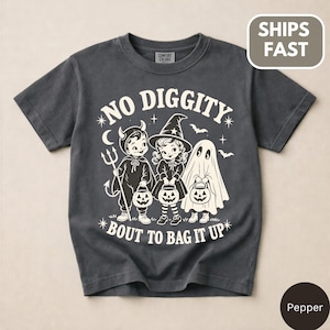 Puede incluir: Una camiseta de manga corta gris carbón con un gráfico color crema de tres niños con disfraces de Halloween: un diablo, una bruja y un fantasma, cada uno con un cubo de calabaza. El texto dice NO DIGGITY BOUT TO BAG IT UP.