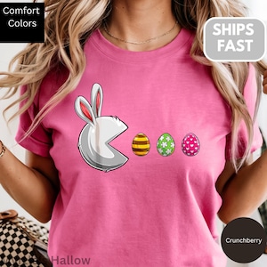May include: Pink t-shirt featuring a Pac-Man-style bunny eating Easter eggs. The bunny is white with pink-lined ears. The eggs are decorated with stripes, flowers, and hearts. The shirt has the words "Comfort Colors" and "Hallow" on it.