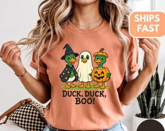 Puede incluir: Camiseta color terracota con un diseño de Halloween. El diseño incluye tres patos de dibujos animados disfrazados de bruja, fantasma y calabaza. El texto "DUCK, DUCK, BOO!" está impreso debajo.