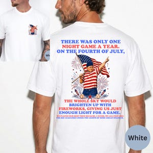 Puede incluir: Camiseta blanca con un diseño patriótico. La parte posterior de la camiseta muestra a un niño sosteniendo una bandera con fuegos artificiales y texto. La parte delantera tiene un pequeño bolsillo con un diseño similar. El texto en la parte posterior dice "There was only one night game a year. On the Fourth of July."