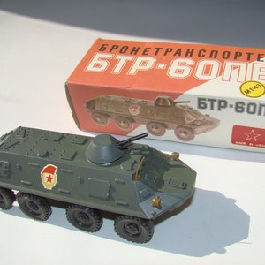 Puede incluir: Un modelo de juguete verde de un vehículo blindado de transporte de personal BTR-60PB, con una estrella roja y una figura de soldado en el lateral. El juguete está en su caja original, que está etiquetada con "Бронетранспортер БТР-60ПБ" y "M 1:43".