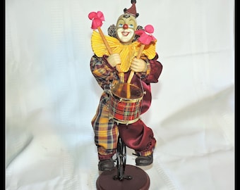 May include: A whimsical clown figurine playing a drum. The clown wears a plaid outfit with a yellow ruffled collar and a small hat. The drum is red plaid, and the figurine stands on a wooden base. The clown holds pink flower-shaped drumsticks.