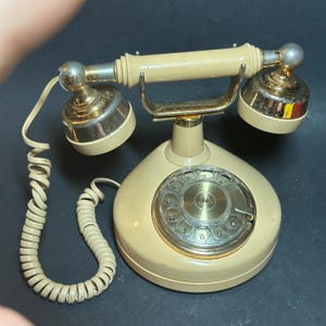 May include: Vintage cream-colored rotary dial telephone with a gold-tone cradle and earpieces. The phone features a circular dial with numbers and a coiled handset cord. The earpieces have a silver finish.