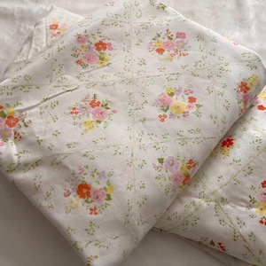 May include: White floral bedsheets with a repeating pattern of colorful flower bouquets. The bouquets include pink, orange, and yellow flowers with green leaves. The sheets are folded, showing the pattern and texture.