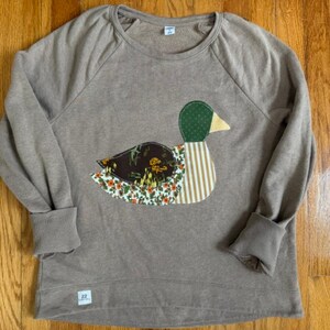 Puede incluir: Una sudadera de manga larga color canela con un gran aplique en forma de pato en la parte delantera. El pato está hecho de varios parches de tela que incluyen lunares verdes, patrones florales marrones y rayas beige. Una etiqueta blanca en el cuello.
