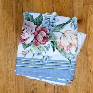 May include: A white fabric sheet featuring a floral pattern with large pink and red roses, pale yellow tulips, and small blue flowers. The bottom edge is defined by a wide light blue border with three horizontal green and white striped lines.