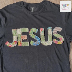 May include: A black crew neck t-shirt featuring the word JESUS appliqued across the chest. The letters are constructed from various colorful floral and patterned fabric patches with visible stitching along the edges of each letter.